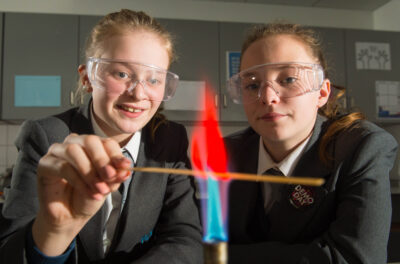 A photo of two high school students wearing safety goggles holding a thin strip of wood over the flame of a Bunsen burner.